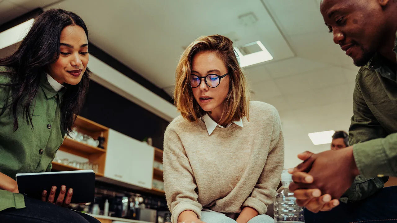 Three people gathered around discussing something; one holds an electronic tablet while others look engaged in conversation within what appears to be an informal meeting space or café.
