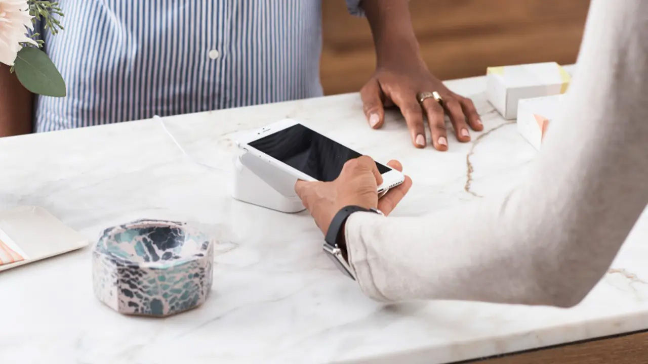 Close-up view showing hands of two individuals making contactless payment using smartphone over point-of-sale terminal placed atop marble countertop.