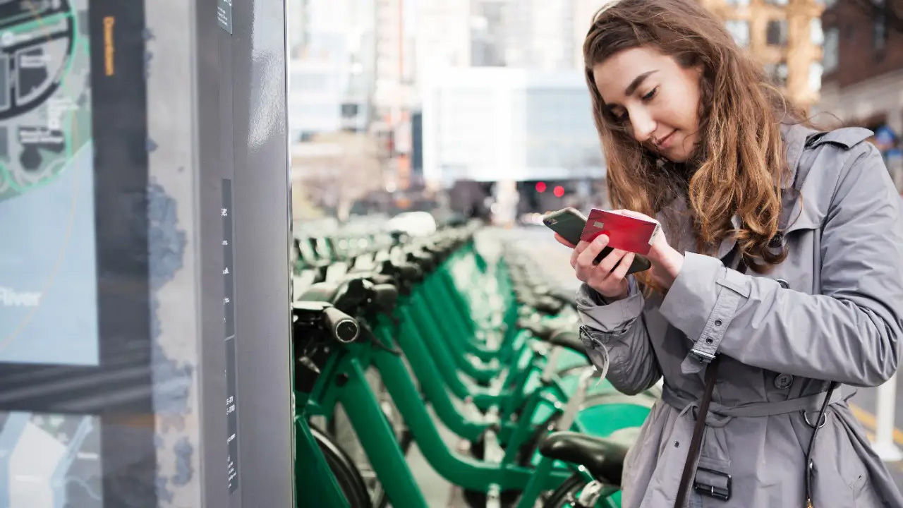 A woman standing next to a bike rental station interacting with a touchscreen to rent an on-demand bike.