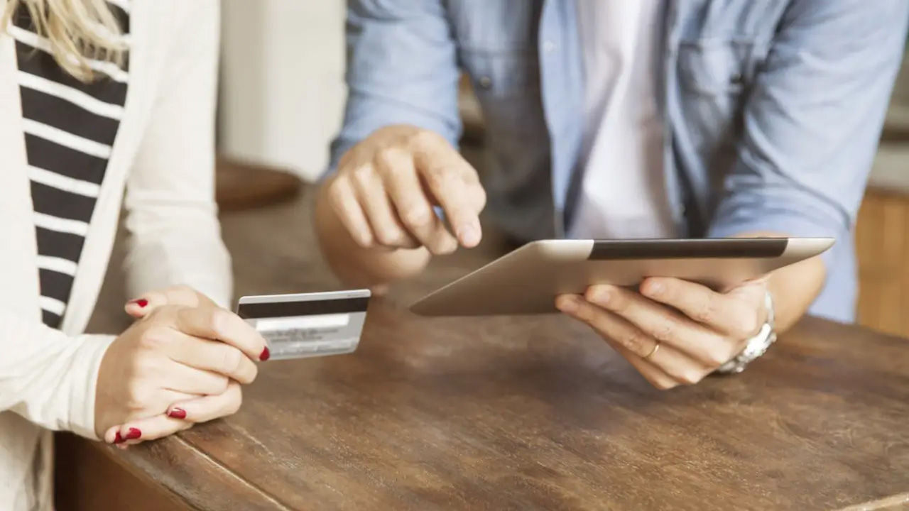 Two people sitting at a table; one holds up a credit card while the other points to something on an electronic tablet they are both looking at.