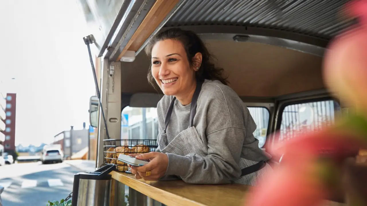 A person smiling while leaning out from inside food truck holding mobile payment device ready for transaction.