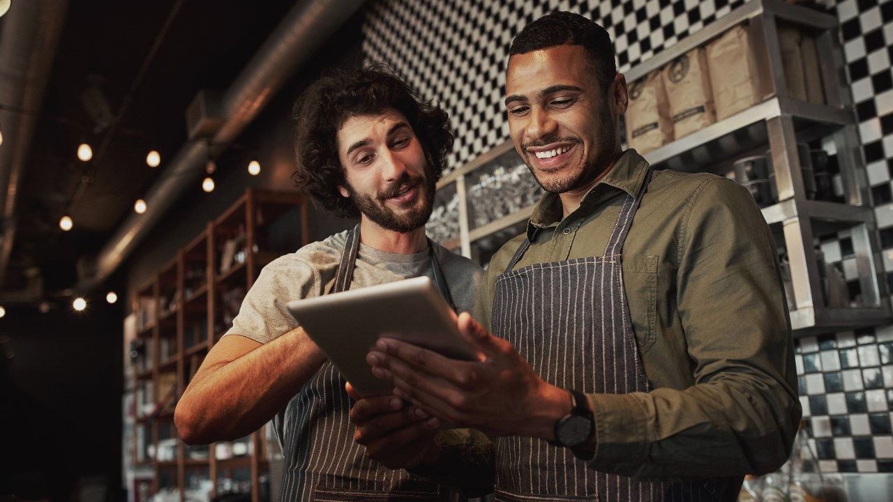 Two men wearing aprons look at a computer tablet together. 