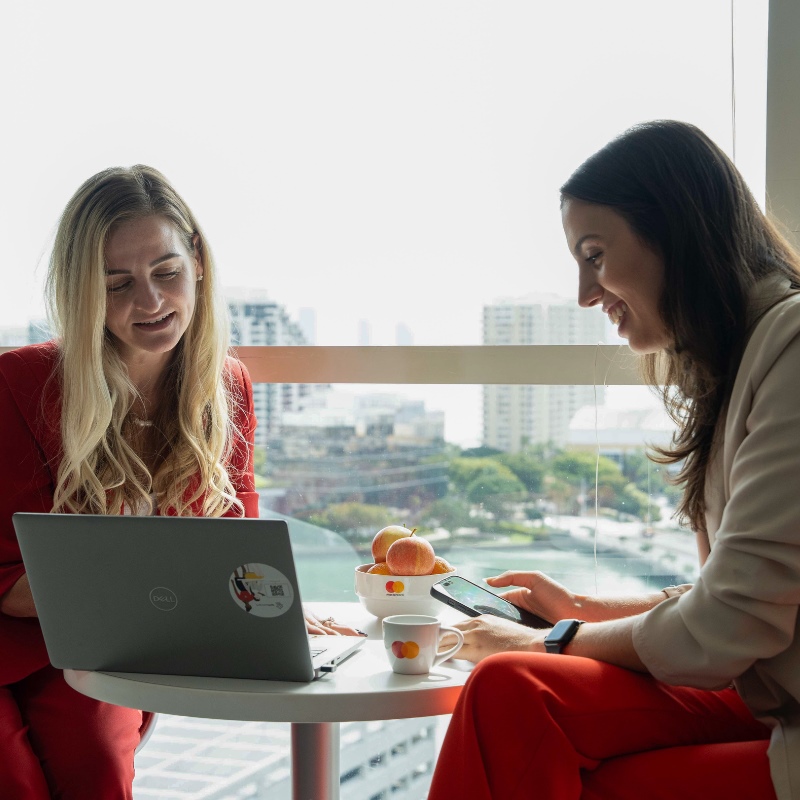 Two employees at Mastercard's Miami office confer over a computer in front of a large window overlooking the skyline. 