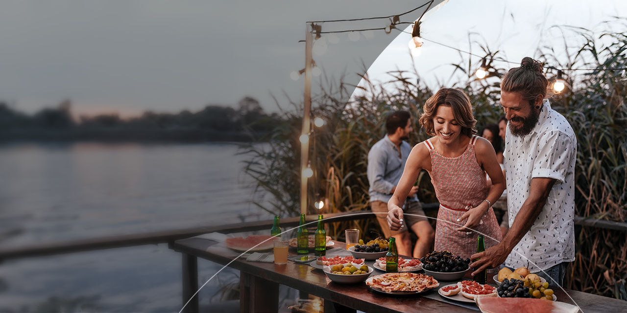 Um casal a sorrir à beira de uma mesa junto ao lago com comida, desfrutando de um encontro alegre com amigos.