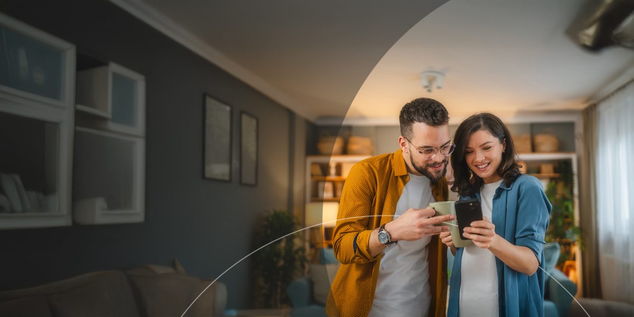 Marido e mulher felizes a beber uma chávena de café enquanto utilizam o telemóvel.