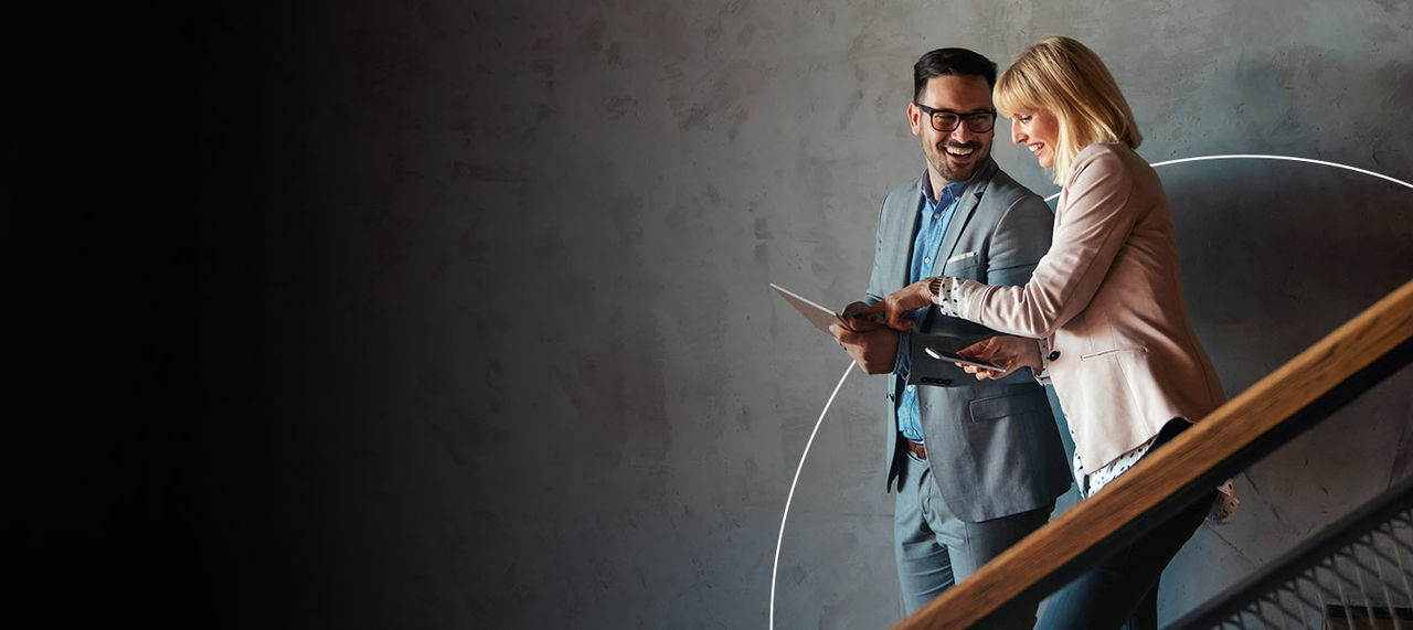 Žena koja se naginje preko svog stola da bi proverila neke detalje sa svog računara u svetlom kancelarijskom prostoru, ukrašenom malim saksijskim biljkama
