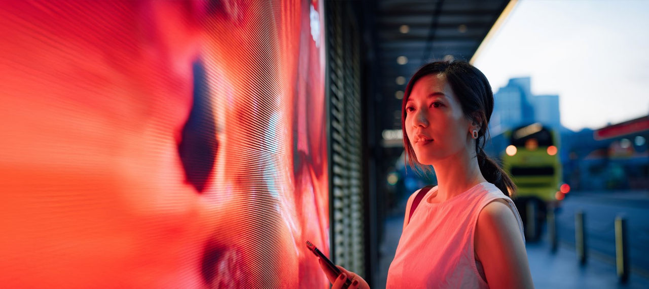 A young woman holds a smartphone and looks at a digital messaging board on a city street. 