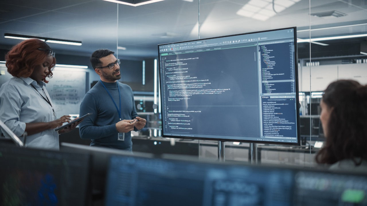 Colleagues look at a large screen with coding on it in an office. 