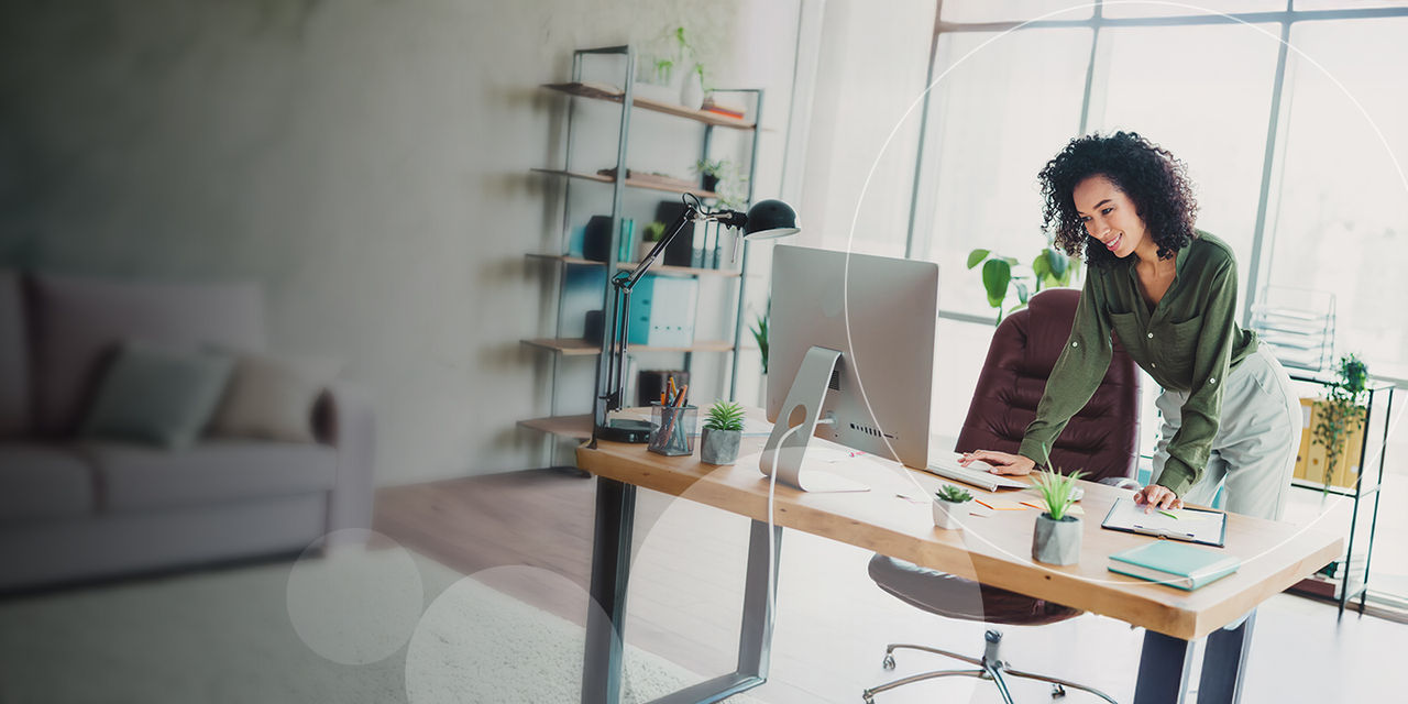 A woman leaning over her desk to check some details from her computer in a bright office space, decorated with small potted plants.