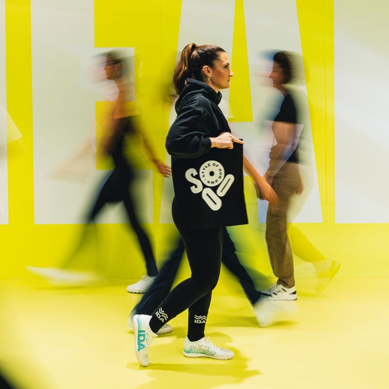 A woman wearing athletic wear and carrying a SOOO bag walks against a yellow background. 