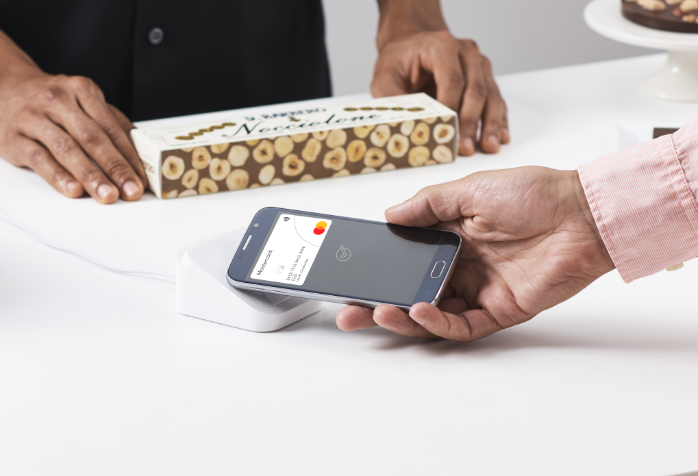 A close-up of a hand holding a phone up to a small white contactless terminal on a counter, with a boxed product and a shop assistant behind.
