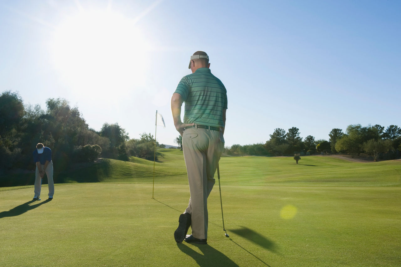 Two golfers on a sunlit green, one standing with a club and the other bending to putt, with a flag and trees in the background.