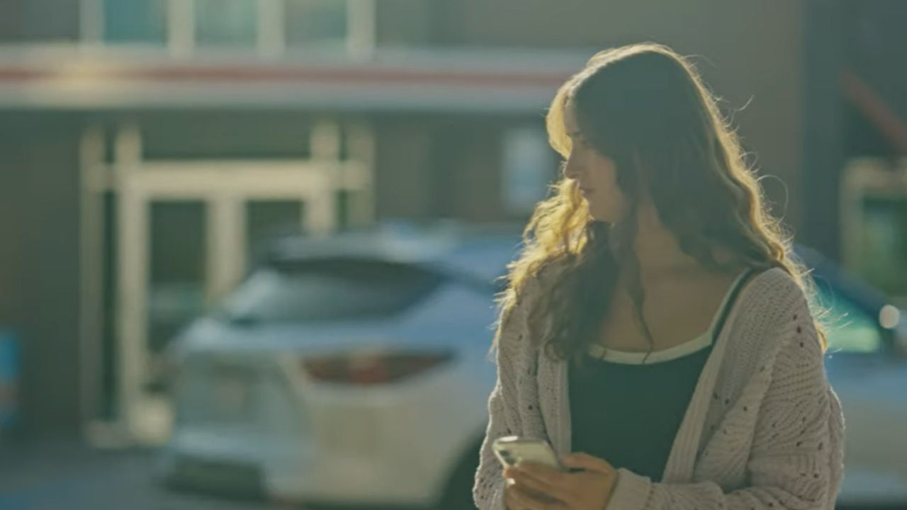 A teenaged girl holds her phone in a parking lot looking worried. 