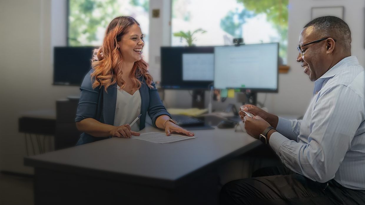 A woman and man are seated at a desk in an office smiling.