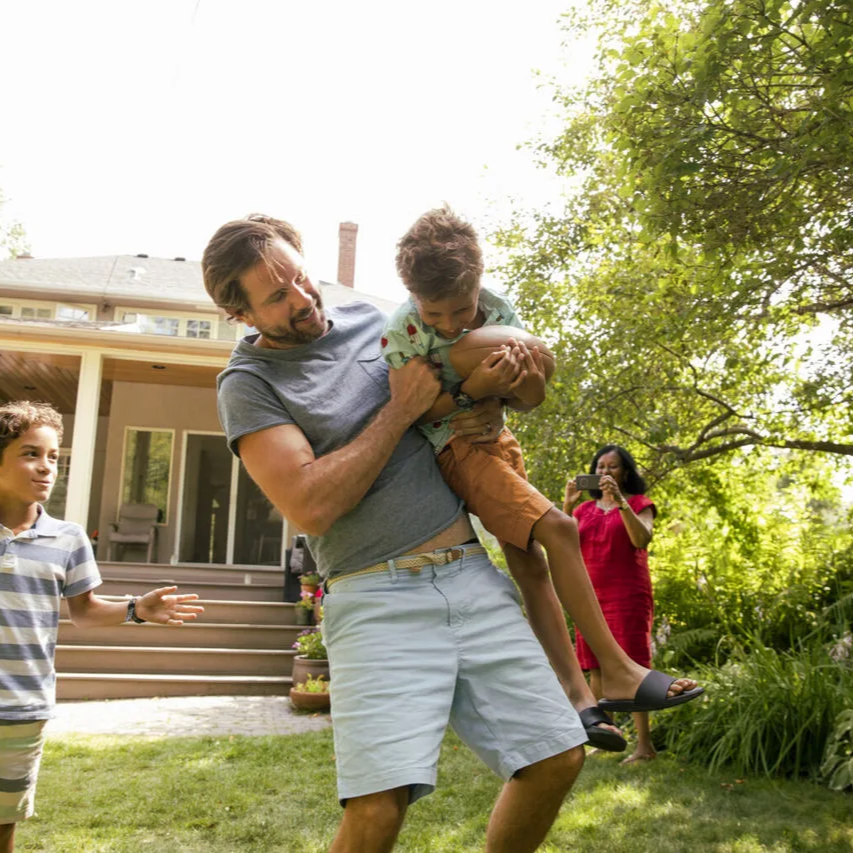 Family playing outside in the front yard while man holds boy with football.