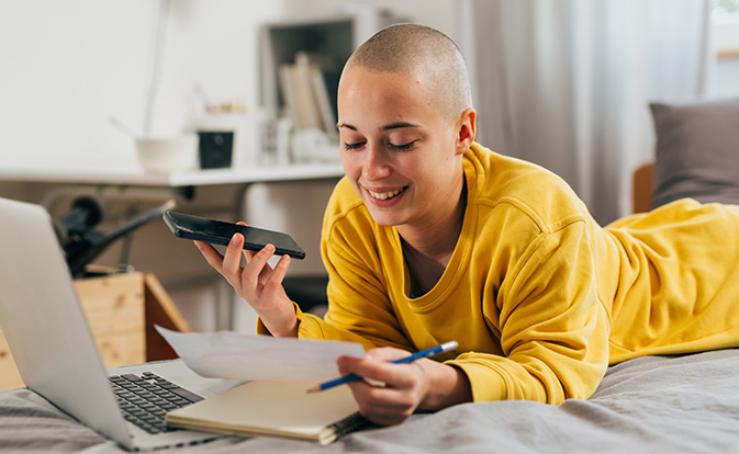 A woman in a yellow sweater relaxes on her bed, using a laptop and a cell phone.