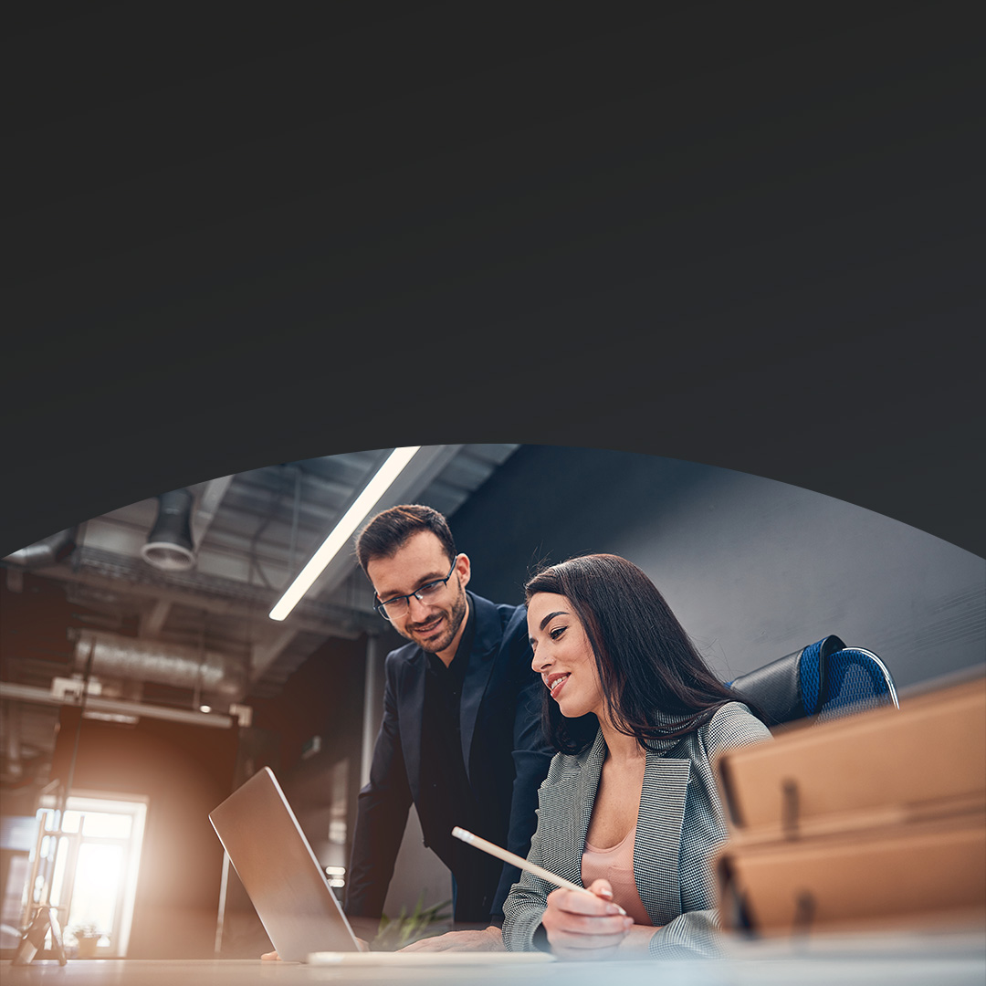 Woman seated at a table shows data on her laptop to a standing male colleague as they discuss.