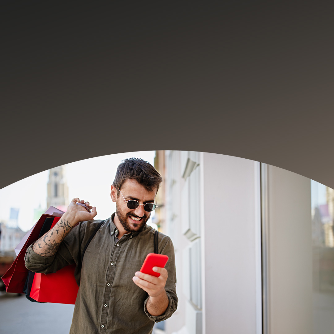 Man smiling as he uses his smartphone, holding shopping bags in his other hand.