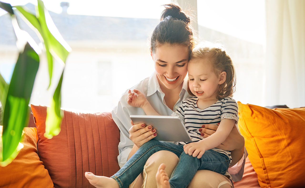 Mother and child smiling while using a tablet together on a couch in a bright room.
