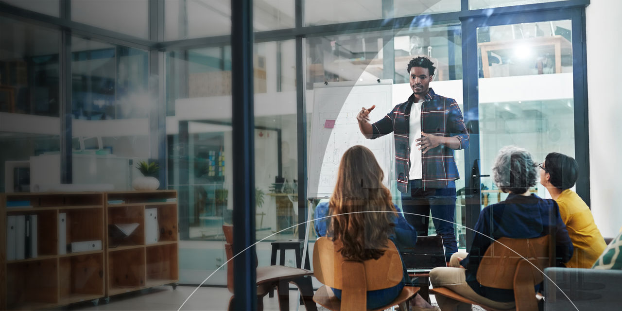 A man stands and gestures while presenting to three seated people in a modern office with glass walls and shelves in the background.