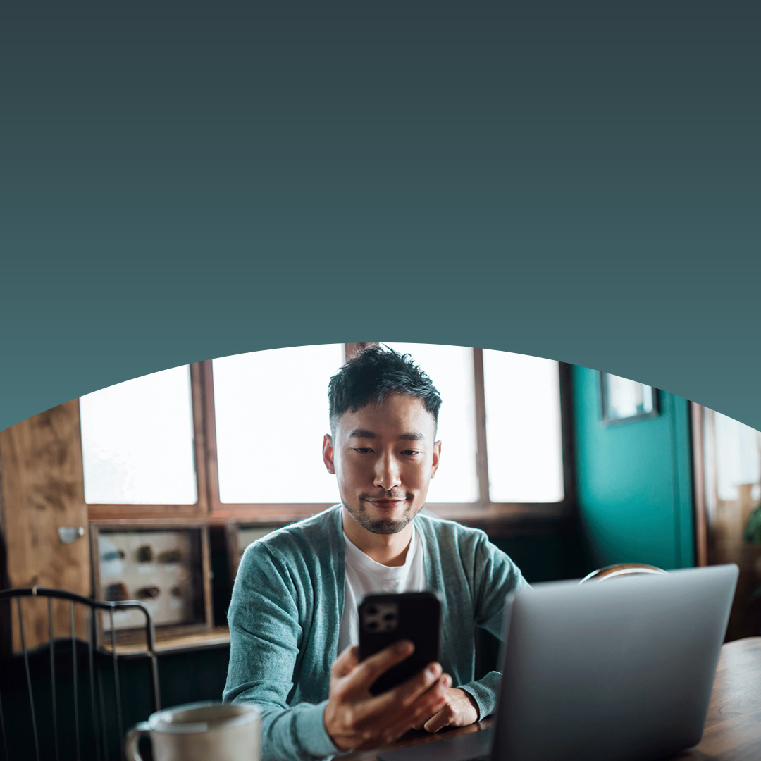 Man working on a laptop while holding a smartphone in his other hand.
