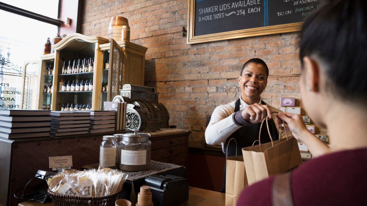A woman shopkeeper hands a bag to a customer. 