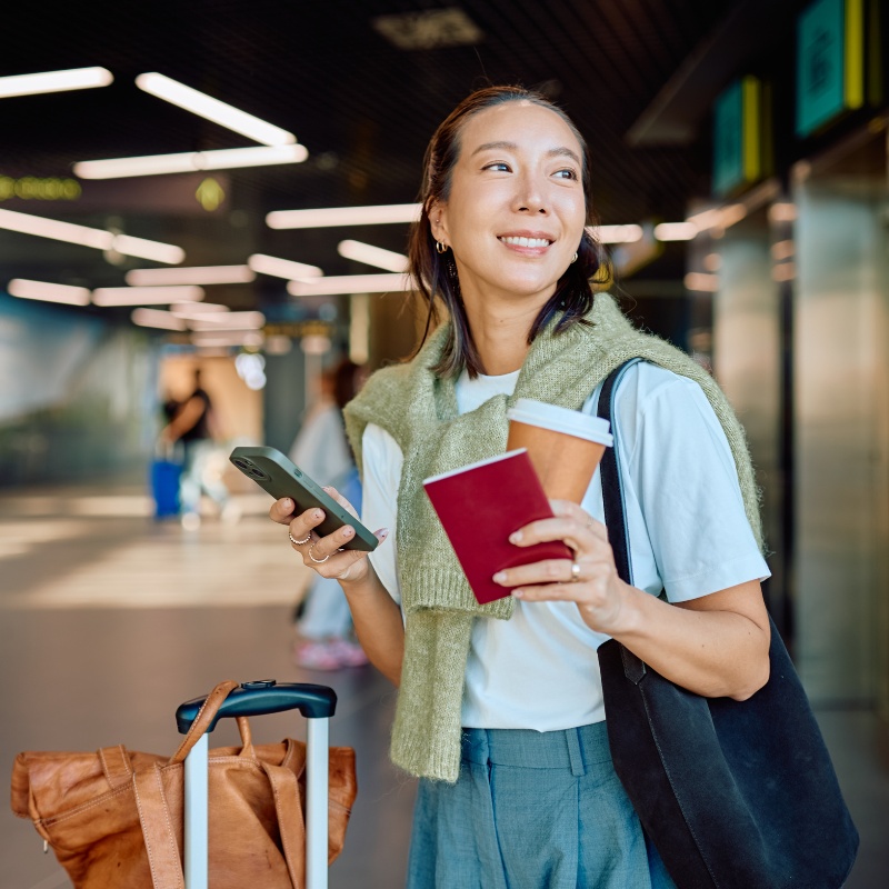 A woman a phone and her passport and a cup of coffee with her suitcase next to her in a transit area. 