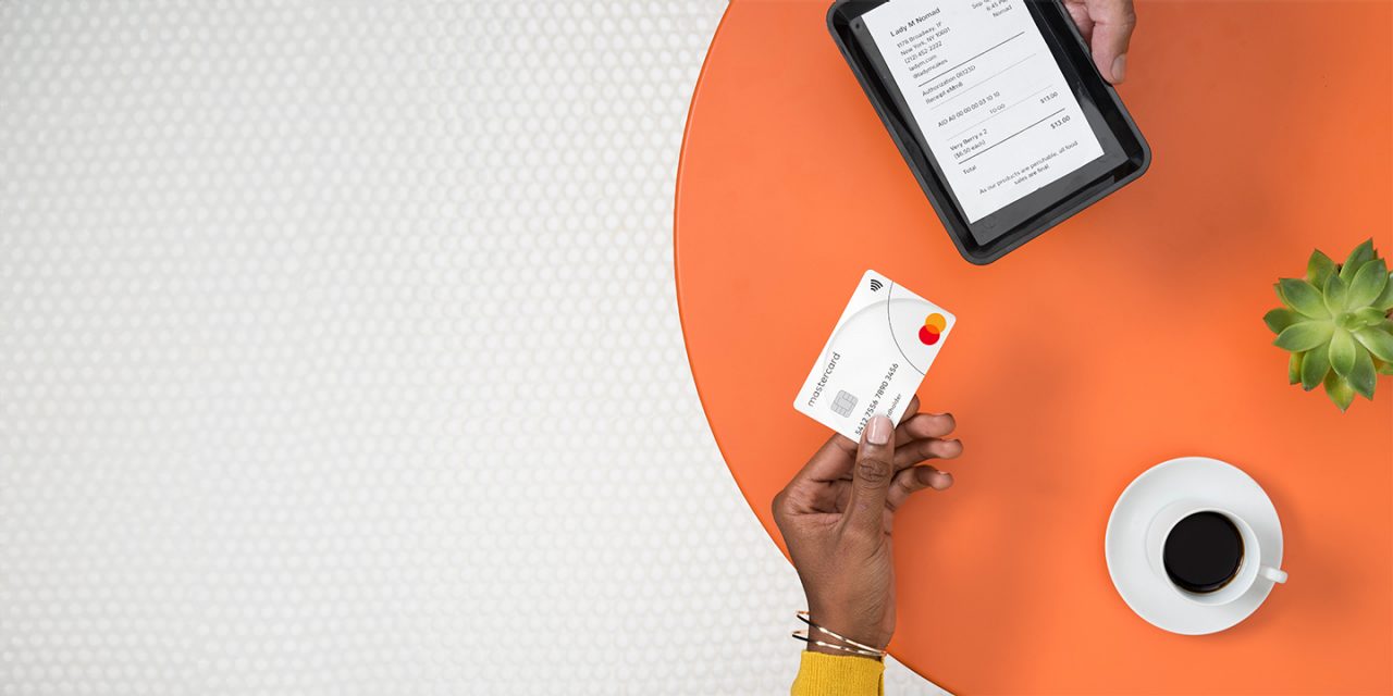 Un consumidor realiza una compra con una tarjeta cripto de Mastercard, mientras que otra mano sostiene una tableta que muestra una pantalla de pago.