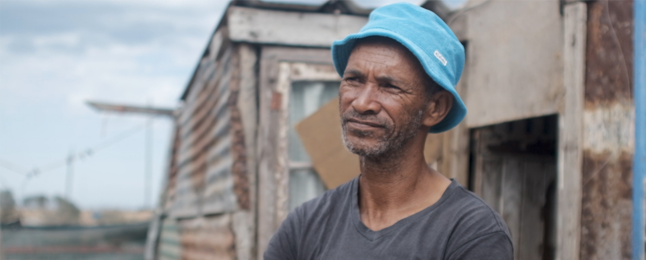 A man in a blue bucket hat and gray T-shirt stands in front of a tin-cladded building at the docks.